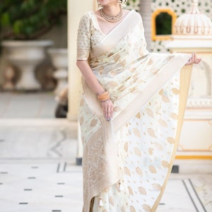 May include: A woman wearing a white and gold saree with a floral pattern. The saree has a gold border and a white tassel.