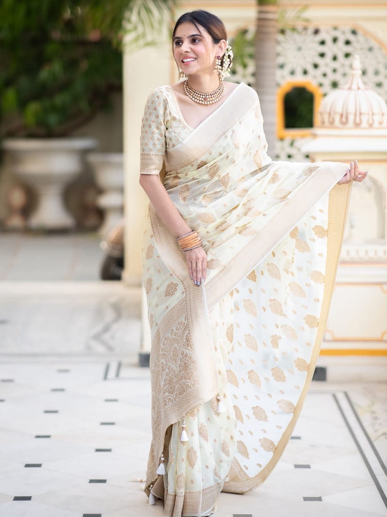 May include: A woman wearing a white and gold saree with a floral pattern. The saree has a gold border and a white tassel.