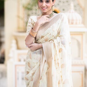 May include: A woman wearing a white and gold saree with a floral pattern. She is smiling and looking at the camera. The saree has a gold border and a gold and white floral design.