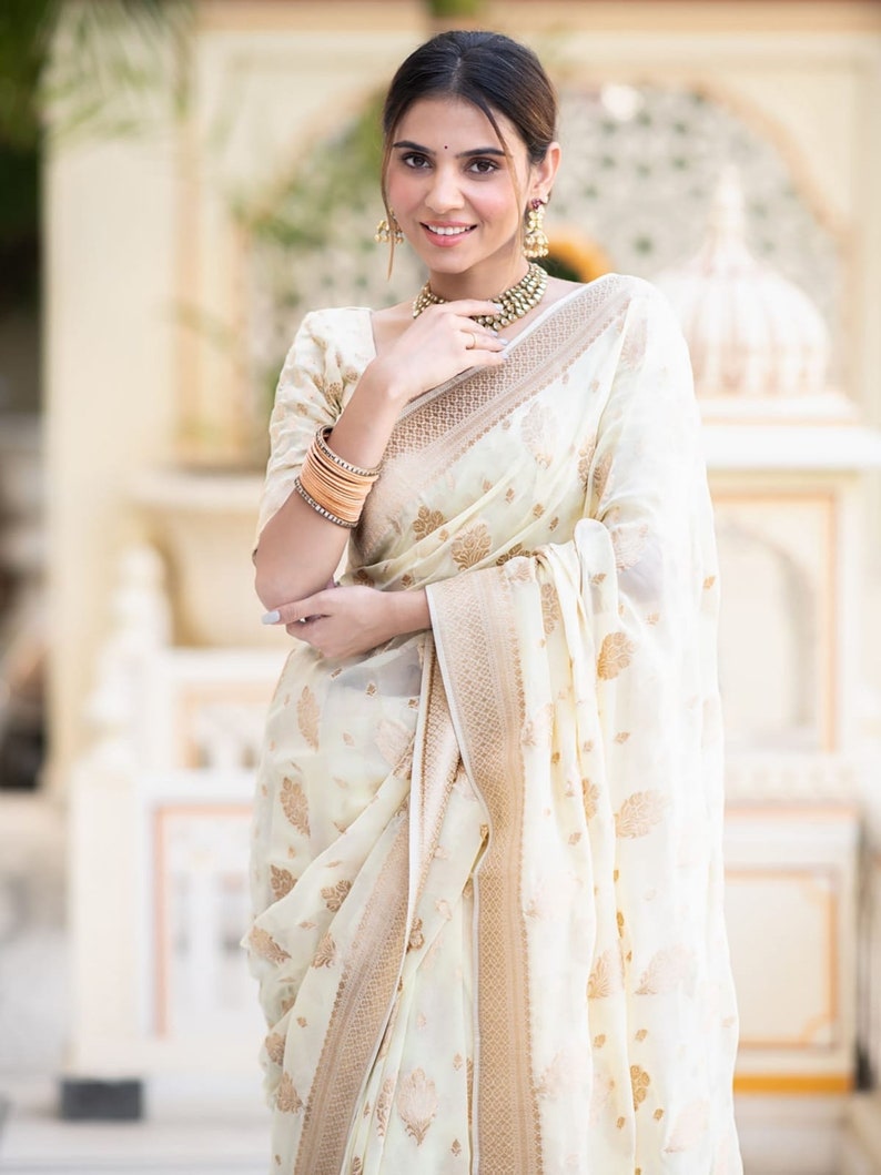 May include: A woman wearing a white and gold saree with a floral pattern. She is smiling and looking at the camera. The saree has a gold border and a gold and white floral design.