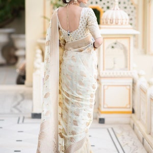 May include: A woman wearing a white and gold saree with a floral pattern. The saree has a gold border and a gold tassel. The woman is standing in front of a white wall.