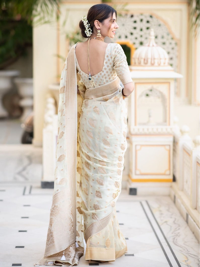 May include: A woman wearing a white and gold saree with a floral pattern. The saree has a gold border and a gold tassel. The woman is standing in front of a white wall.