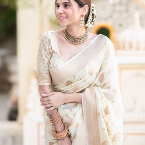 May include: A woman wearing a white and gold saree with a gold necklace and earrings. She is standing in front of a white wall.
