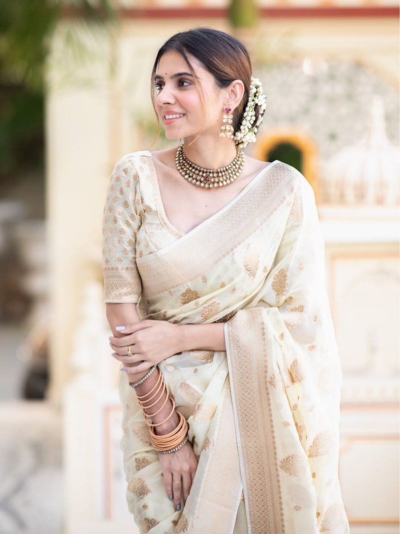 May include: A woman wearing a white and gold saree with a gold necklace and earrings. She is standing in front of a white wall.