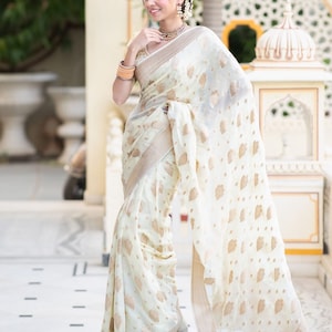 May include: A woman wearing a white and gold saree with a floral pattern. The saree is draped over her shoulders and waist, and she is standing in front of a white building.