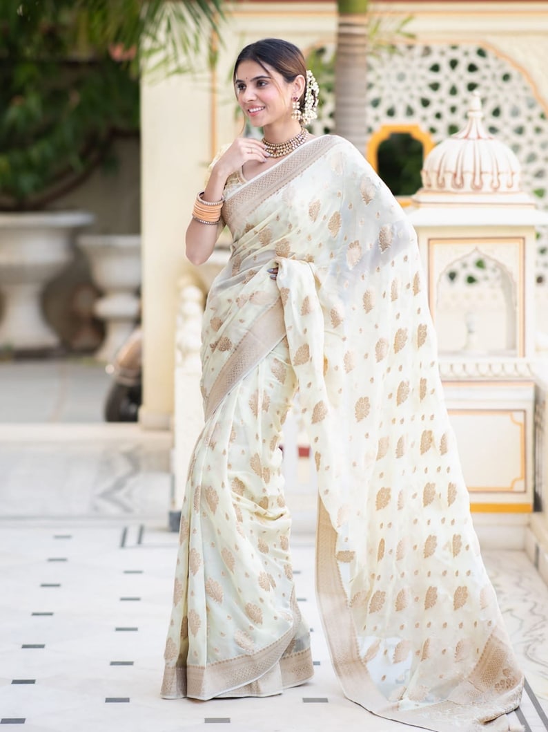 May include: A woman wearing a white and gold saree with a floral pattern. The saree is draped over her shoulders and waist, and she is standing in front of a white building.