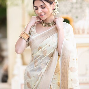 May include: A woman wearing a white and gold saree with a floral pattern. She is wearing a pearl necklace and earrings. The saree has a gold border.