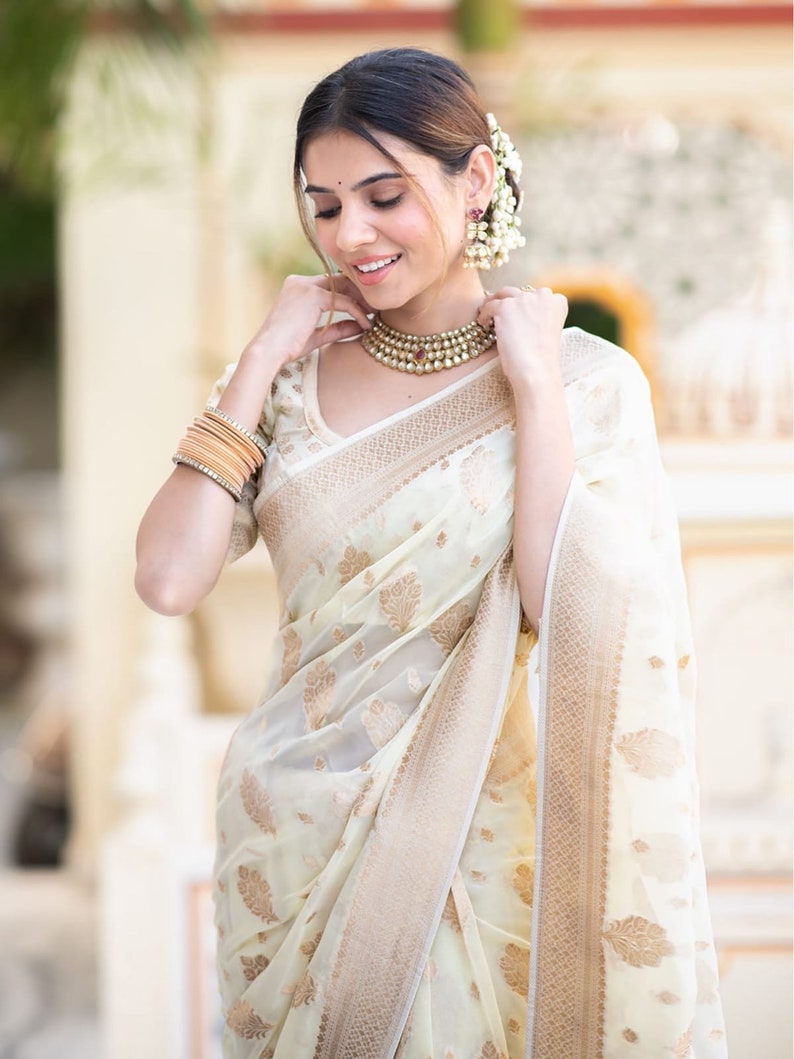 May include: A woman wearing a white and gold saree with a floral pattern. She is wearing a pearl necklace and earrings. The saree has a gold border.