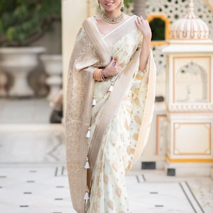 May include: A woman wearing a white and gold saree with a floral pattern. The saree has a gold border and tassels. The woman is standing in front of a white building with a decorative archway.