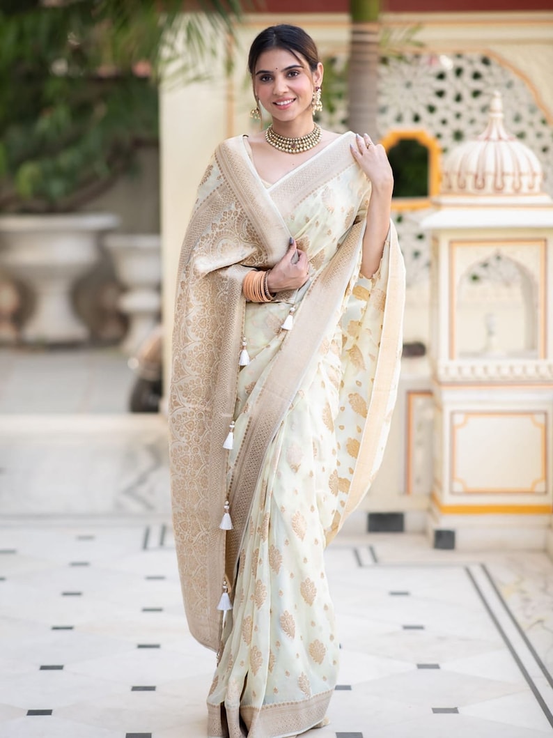 May include: A woman wearing a white and gold saree with a floral pattern. The saree has a gold border and tassels. The woman is standing in front of a white building with a decorative archway.
