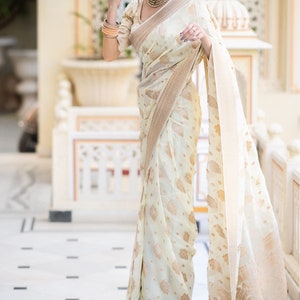 May include: A woman wearing a white and gold saree with a floral pattern. The saree has a gold border and is draped over a white blouse.