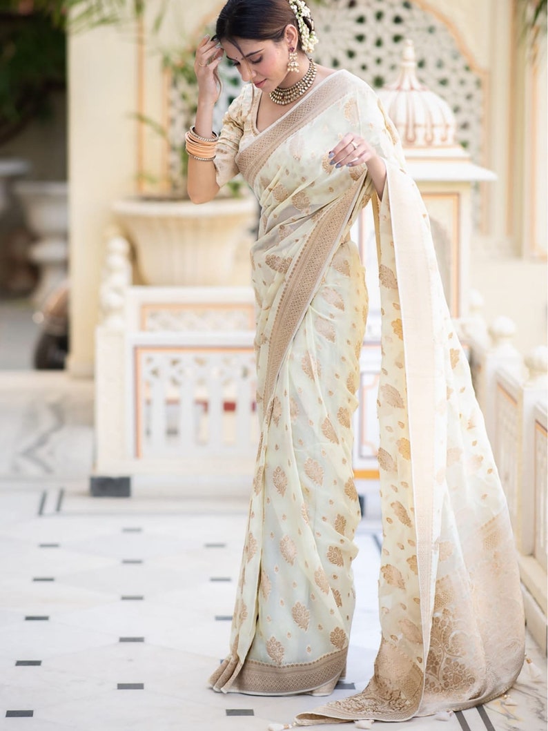 May include: A woman wearing a white and gold saree with a floral pattern. The saree has a gold border and is draped over a white blouse.