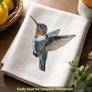 May include: A white kitchen towel featuring a detailed embroidered hummingbird in flight. The bird displays vibrant shades of blue, orange, and brown thread, capturing the intricate texture of its feathers and wings against the plain fabric.