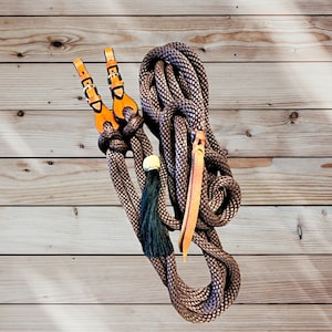 May include: A brown and gray rope with leather accents and a decorative tassel. The rope features two orange leather straps with gold buckles. A long, thin piece of leather is attached to the rope.