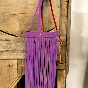 Puede incluir: Un bolso morado con flecos y una correa de cuero marr&oacute;n colgando de un gancho de madera.