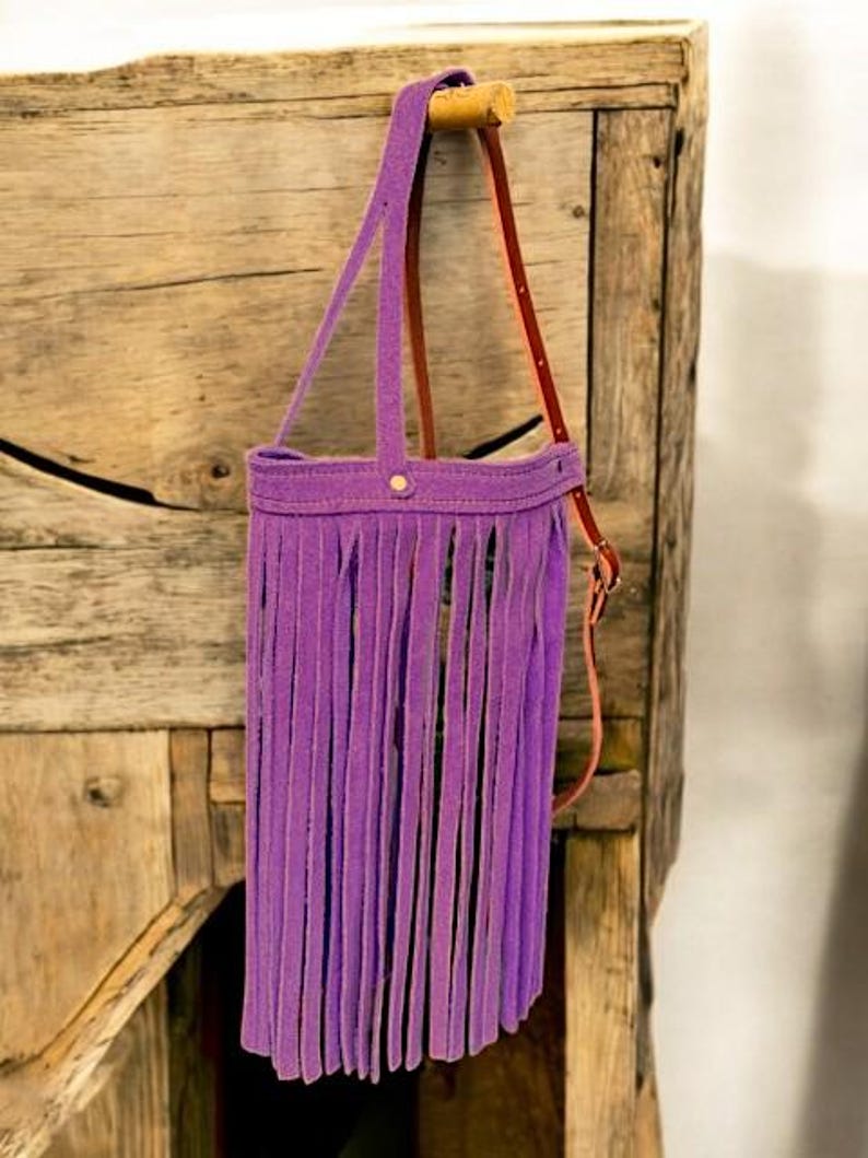 Puede incluir: Un bolso morado con flecos y una correa de cuero marr&oacute;n colgando de un gancho de madera.
