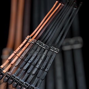 May include: A collection of brown and black leather camera straps with metal buckles. The straps are arranged in a fan shape, with the brown straps on top and the black straps on the bottom.