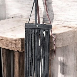 Puede incluir: Un bolso de cuero negro con un detalle de flecos y una correa de cuero marr&oacute;n. El bolso est&aacute; colgado en una superficie de madera.