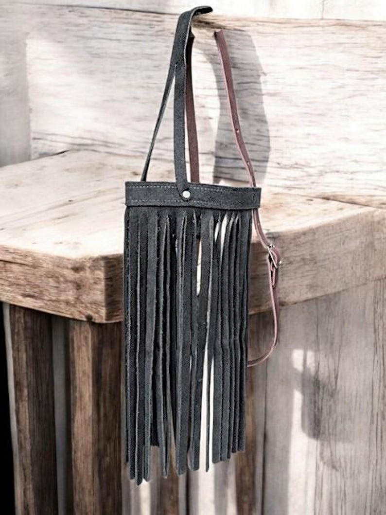 Puede incluir: Un bolso de cuero negro con un detalle de flecos y una correa de cuero marr&oacute;n. El bolso est&aacute; colgado en una superficie de madera.