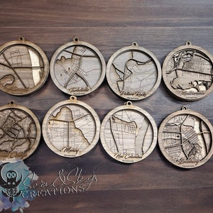 May include: Eight wooden ornaments featuring laser-cut maps of different towns in Pennsylvania. Each ornament is round and has a brown wood finish. The towns featured are: Alexandria, Turbotville, Picture Rocks, Montoursville, New Columbia, Muncy, Lewisburg, and Jersey Shore.