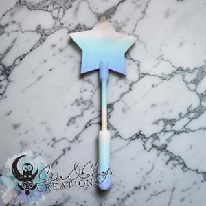 Puede incluir: Una varita mágica en forma de estrella con un degradado de blanco, azul y azul claro. El mango tiene un degradado a juego. La varita está sobre una superficie de mármol. El texto "Sea & Sky Creations" está en la esquina inferior izquierda.