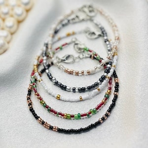 May include: A collection of delicate beaded bracelets in various colours, including black, white, and gold. The bracelets feature small, colourful beads and silver clasps. The bracelets are displayed on a white surface, showcasing their intricate designs.