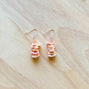 May include: A pair of dangle earrings with a stack of small, peach-colored shell pieces hanging from silver hooks.