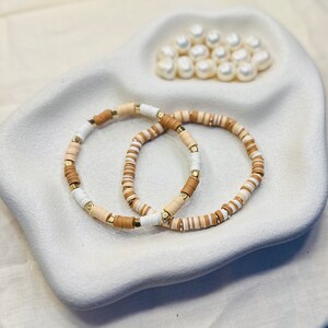 May include: Two bracelets made of white, brown, and gold beads. The bracelets are stacked on top of each other.