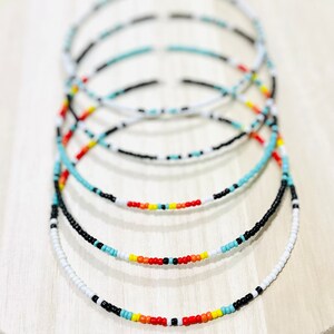 May include: A set of five beaded necklaces in black, white, turquoise, orange, and yellow. The necklaces are layered on top of each other, creating a colourful and eye-catching display.