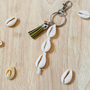 May include: A white shell keychain with a green and brown tassel charm. The keychain is made of cowrie shells strung together with white string and attached to a silver key ring.
