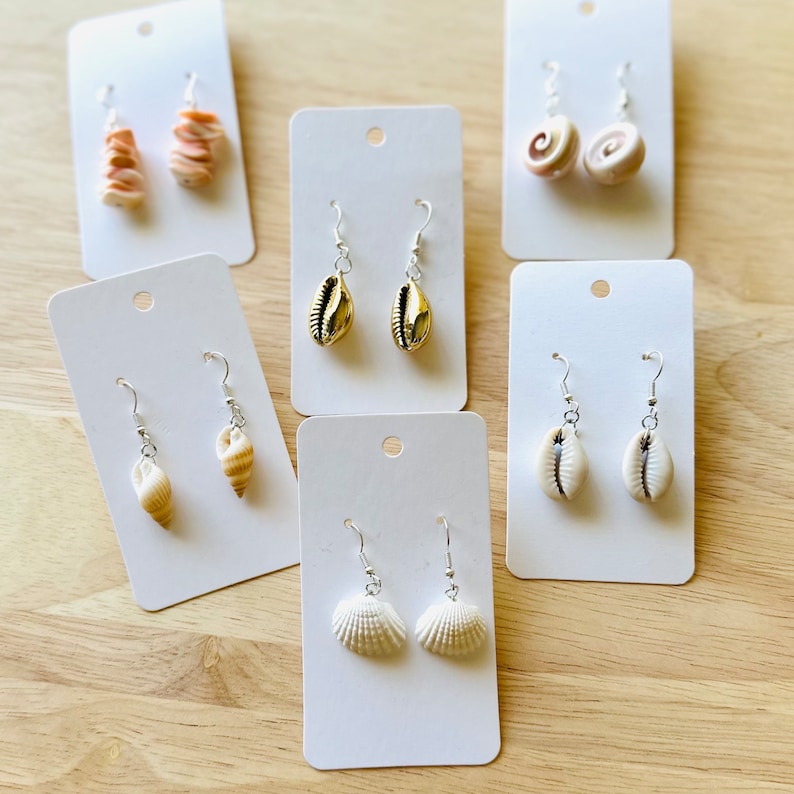 May include: Six pairs of earrings on white cards, each pair featuring a different shell design. The earrings are made of gold, white, and brown shells.