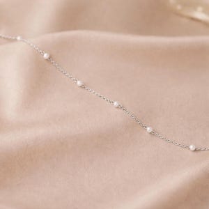 May include: A delicate silver necklace with small, evenly spaced pearls. The necklace is shown against a soft, neutral-toned background, highlighting its elegant simplicity. The chain is thin and the pearls are round.