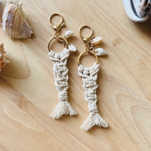 May include: Two white macrame keychains with gold rings and small seashells attached. The keychains are shaped like mermaid tails.