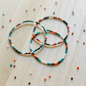 May include: Three beaded bracelets in black, white, orange, and turquoise. The bracelets are layered on a light wood surface.