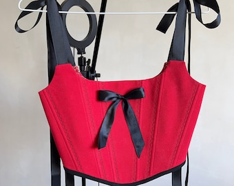 May include: A red corset top with black ribbon straps and a bow. The corset features vertical stitching and a lace-up back. The top is hanging on a white hanger.