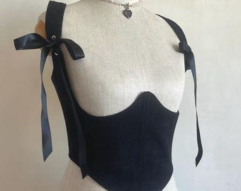 May include: Black corset top with shoulder straps tied with black satin bows. The bustier is displayed on a mannequin. A pink beaded necklace with a heart pendant is around the mannequin's neck. Fashion apparel.