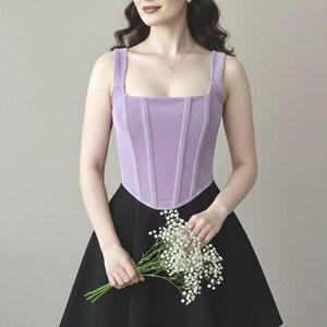 May include: A lavender-colored corset top with a square neckline and vertical ribbing. The top is paired with a black skater skirt. The person is holding a bouquet of white flowers.