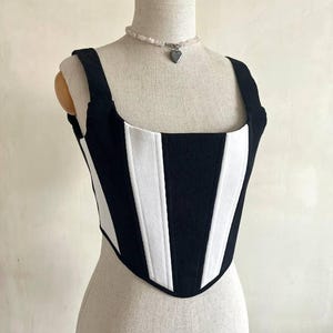 May include: A black and white striped corset top with thick straps. The top is displayed on a mannequin. A beaded necklace with a heart pendant is around the mannequin's neck. The corset has a square neckline and a fitted silhouette.
