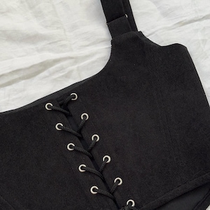 May include: Black corset top with a corded lace-up front and adjustable shoulder strap. The top features silver eyelets and a buckle. The fabric has a ribbed texture. The garment is designed with a fitted silhouette.
