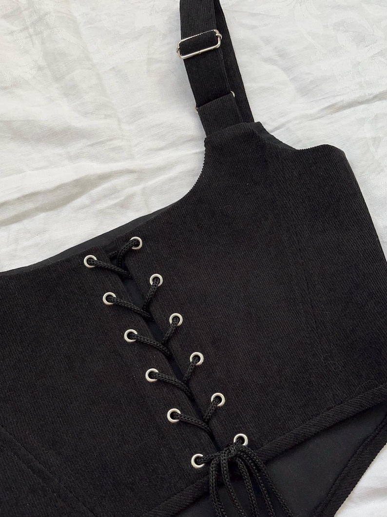 May include: Black corset top with a corded lace-up front and adjustable shoulder strap. The top features silver eyelets and a buckle. The fabric has a ribbed texture. The garment is designed with a fitted silhouette.