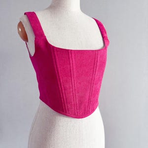 May include: A bright fuchsia corduroy corset top with a square neckline and wide shoulder straps. The structured bodice features vertical seams for a tailored fit. The top is displayed on a mannequin.