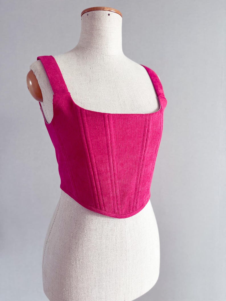 May include: A bright fuchsia corduroy corset top with a square neckline and wide shoulder straps. The structured bodice features vertical seams for a tailored fit. The top is displayed on a mannequin.