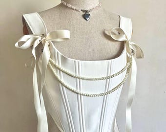 May include: A white corset top featuring vertical boning, wide shoulder straps tied with satin ribbons, and two draped strands of white faux pearls across the front. The garment is displayed on a beige mannequin wearing a heart pendant necklace.