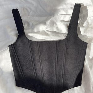 May include: A black corset top with wide shoulder straps and vertical ribbing. The top features a square neckline and a lace-up detail at the front. The garment is made of a ribbed fabric.