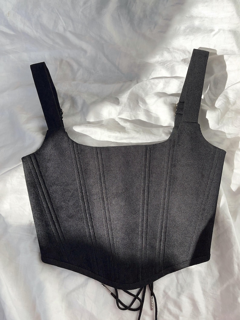 May include: A black corset top with wide shoulder straps and vertical ribbing. The top features a square neckline and a lace-up detail at the front. The garment is made of a ribbed fabric.