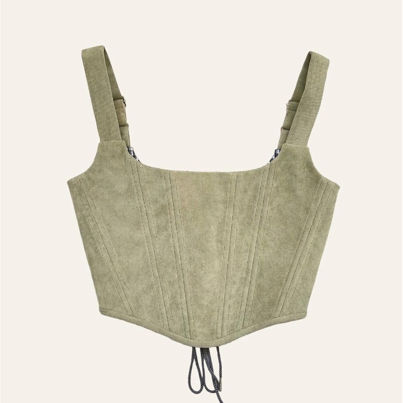 May include: A sage green corset top with adjustable straps and a square neckline. The structured bodice has vertical seam detailing and a lace-up closure. The straps are attached with metal hardware.