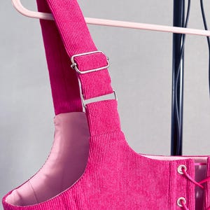 May include: Close-up of a vibrant pink corduroy garment featuring adjustable straps and silver metal hardware. The garment has a laced front and is suspended from a pink clothes hanger.