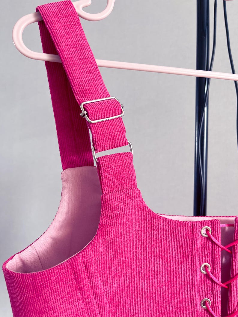 May include: Close-up of a vibrant pink corduroy garment featuring adjustable straps and silver metal hardware. The garment has a laced front and is suspended from a pink clothes hanger.