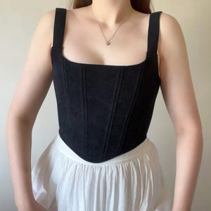 May include: Black corduroy corset top with wide shoulder straps and a square neckline. The top is paired with a white skirt. The corset has vertical seams for a structured fit. A silver necklace is visible.