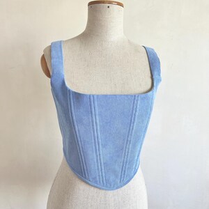 May include: A pale blue corduroy corset top with a square neckline and broad straps. The bodice has vertical seams and a curved hem. The garment is displayed on a mannequin.